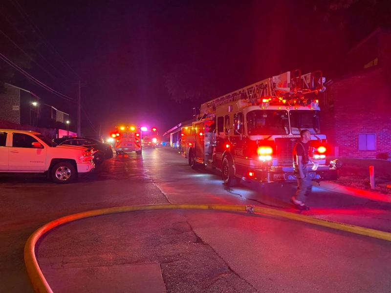 A fire truck with its lights on and hose running from it is parked near the scene of a fire at night. 