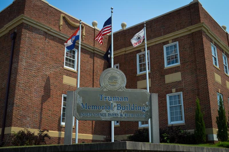 Truman Memorial Building from corner with flag poles behind monument sign and in front of historic red brick building.